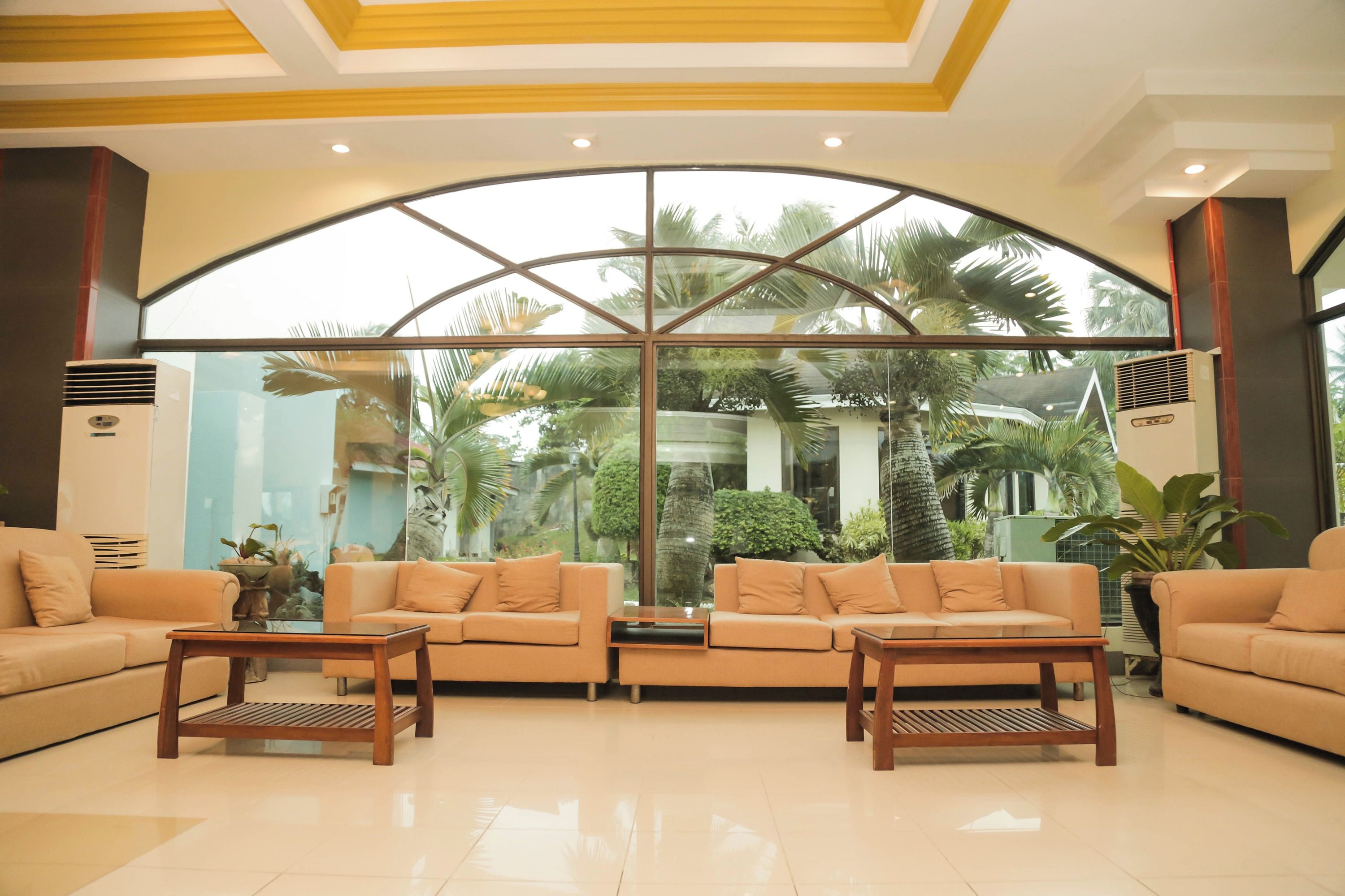 Eine geräumige Lobby mit zwei Sofas und einem Couchtisch, große Fenster mit Blick auf Palmen und Grünflächen, sowie ein modernes Decken-Design.