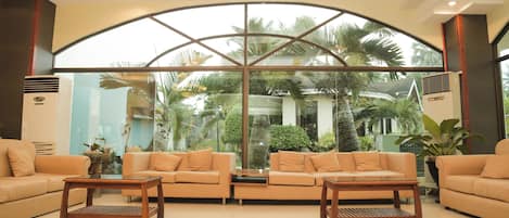 Un vestíbulo espacioso con dos sofás y una mesa de centro, amplias ventanas que ofrecen vistas a palmeras y follaje, y un diseño de techo moderno.