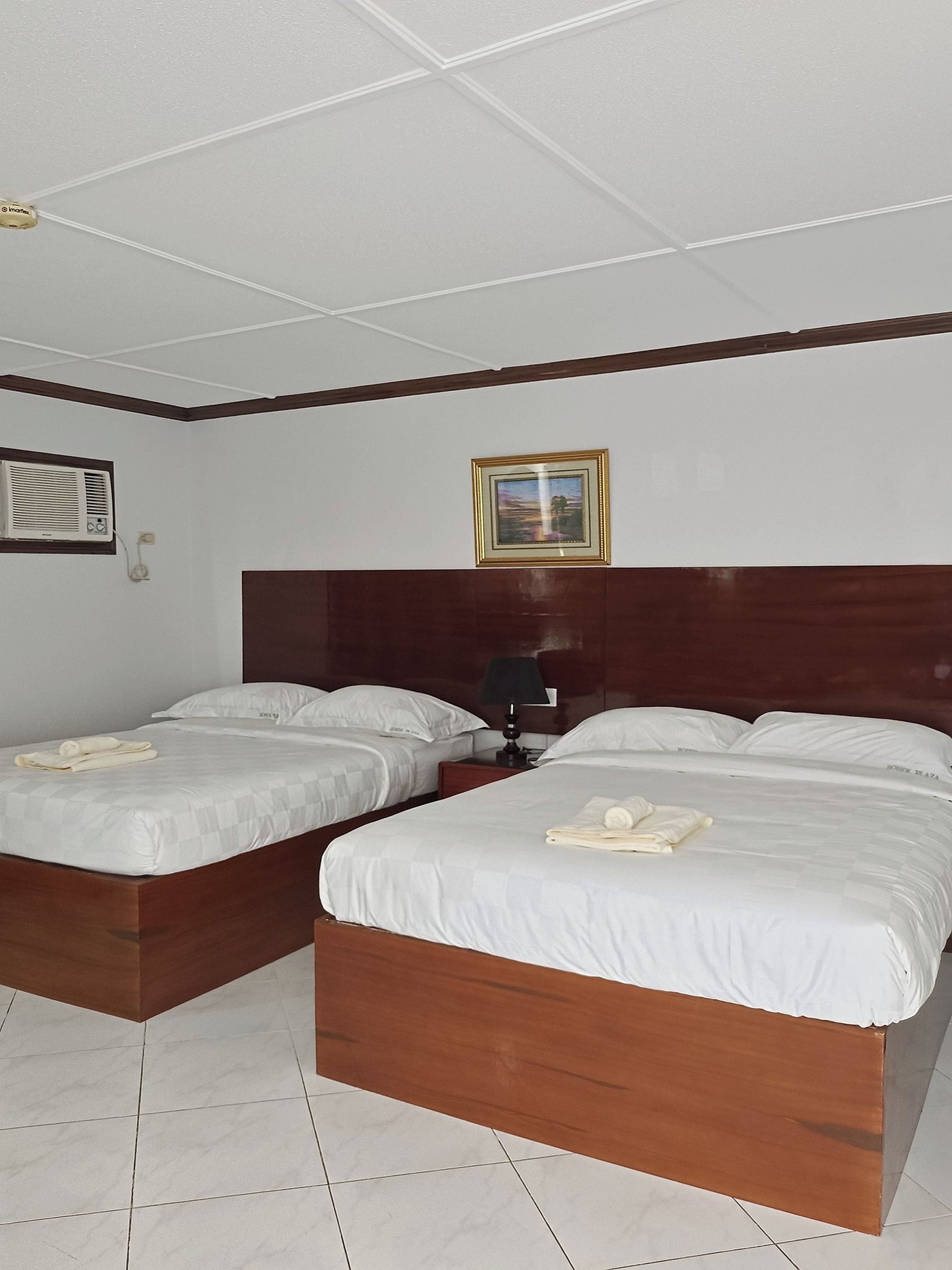 A hotel room with two beds, a wooden headboard, a framed picture, and a bedside table with a lamp.