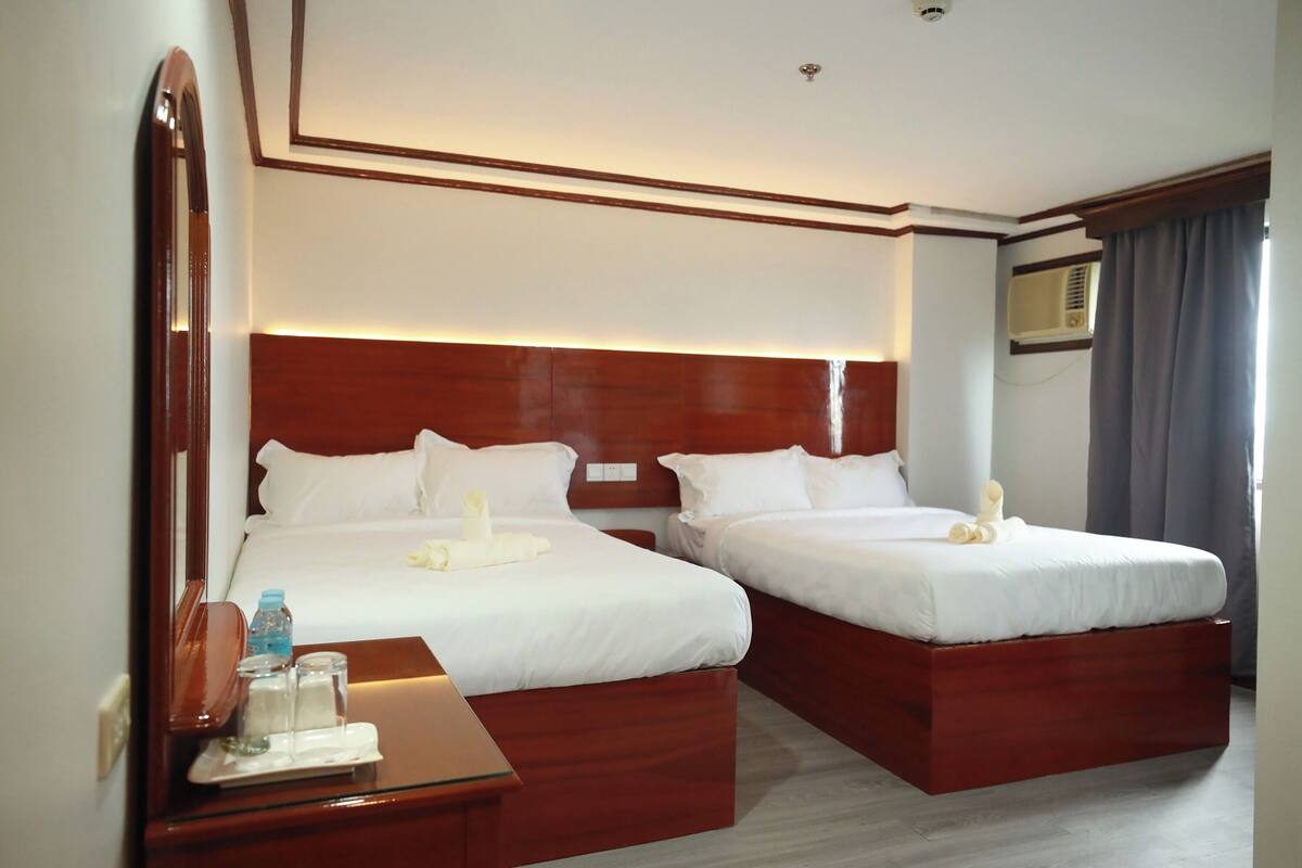 A hotel room with two beds, a wooden headboard, a bedside table with a lamp, and a mirror.