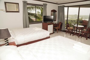 Habitación de hotel con dos camas, un escritorio con televisor, un balcón con sillas y vistas al paisaje.