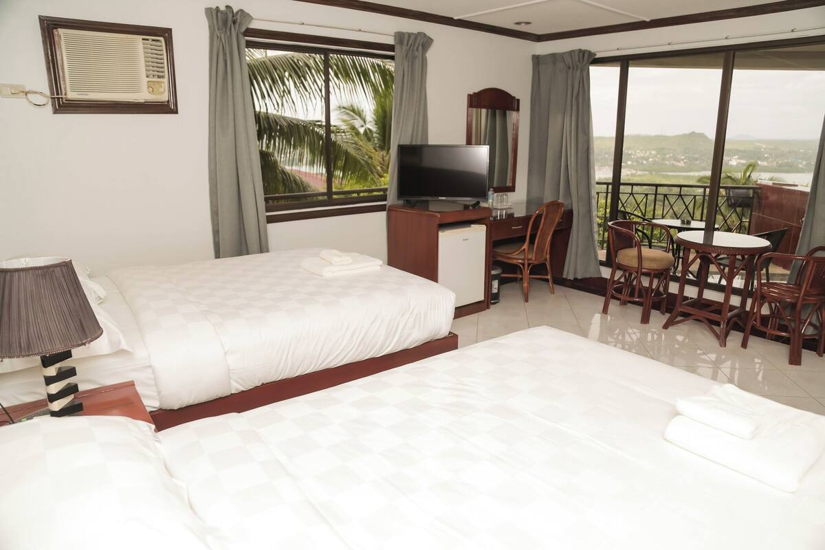 A hotel room with two beds, a desk with a TV, a balcony with chairs, and a view of the landscape.