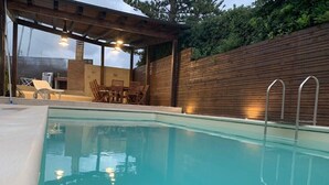 Una piscina con terrazza in legno, zona salotto esterna e patio coperto.