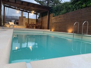 A swimming pool with a wooden deck area, outdoor seating, and a covered patio.