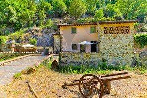A stone building with a tiled roof, a small bridge, and a rustic outdoor area with a wheelbarrow. - Villa Gabriella Chianti Toscana (Greve in Chianti)