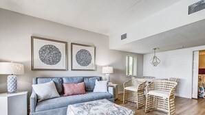 Un salon comprenant un canapé, un pouf et deux fauteuils en osier. Deux œuvres d’art encadrées ornent le mur, une lampe se trouve sur une table d’appoint et un lustre illumine la pièce.