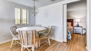 Un coin repas avec une table ronde et quatre chaises en osier, un lustre et une fenêtre avec des stores.
