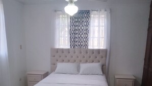 Quarto com cama grande, duas mesas de cabeceira, ventilador de teto e janela com cortinas estampadas.