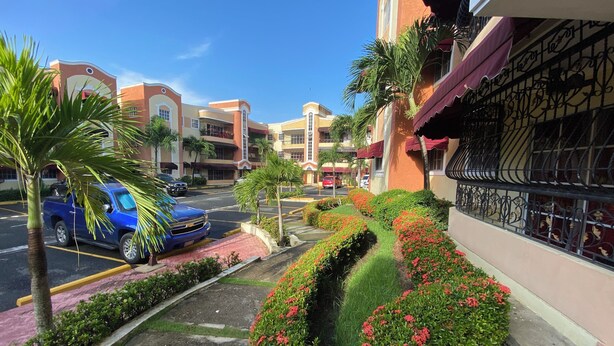 Um estacionamento com uma caminhonete azul, um edifício com varandas e um jardim bem cuidado com flores vermelhas.