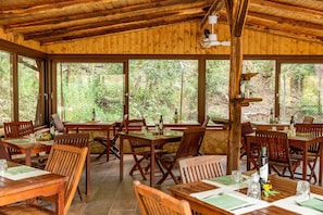 Een houten interieur met tafels en stoelen, grote ramen en uitzicht op bomen.