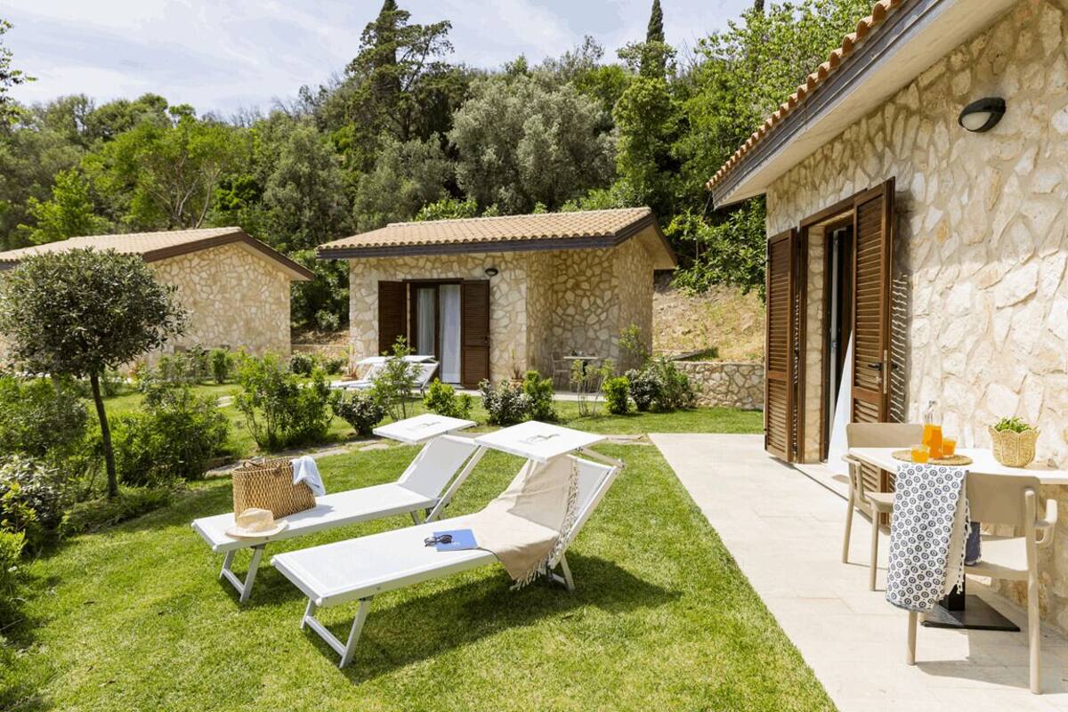 Una villa de piedra con dos zonas de estar, una con sillas blancas y la otra con una mesa y sillas, rodeada de un jardín cuidado.