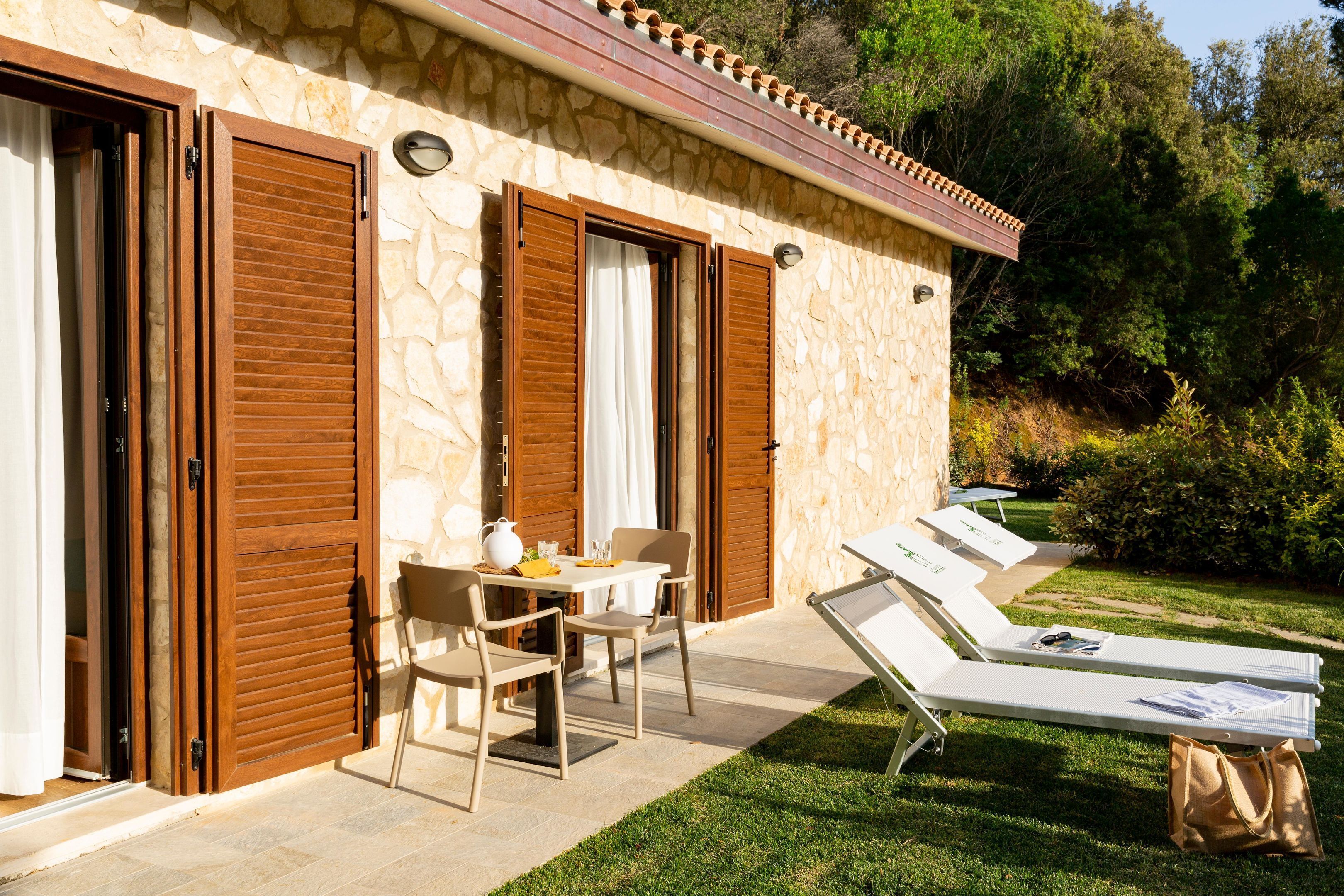 A stone building with wooden shutters, a table with chairs, and sun loungers on a patio.