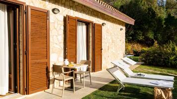 A stone building with wooden shutters, a table with chairs, and sun loungers on a patio.
