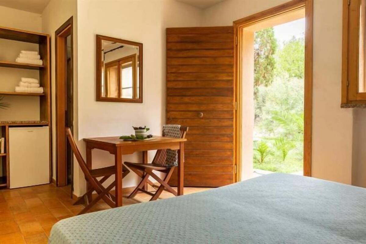 Un dormitorio con cama, escritorio con silla, un refrigerador pequeño y una puerta de madera que da a un balcón.