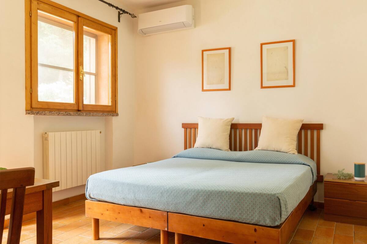 Un dormitorio con cama de madera, ventana con persianas, aire acondicionado, radiador y dos cuadros en la pared.