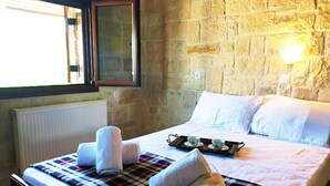 A bedroom with a stone wall, a window with blinds, a radiator, a bed with a plaid blanket, two rolled towels, and a tray with cups and saucers.