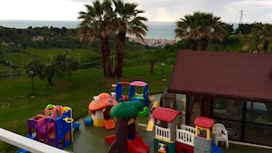 A children's play area with various colorful play structures, a large tree, and a view of the ocean and distant cityscape.