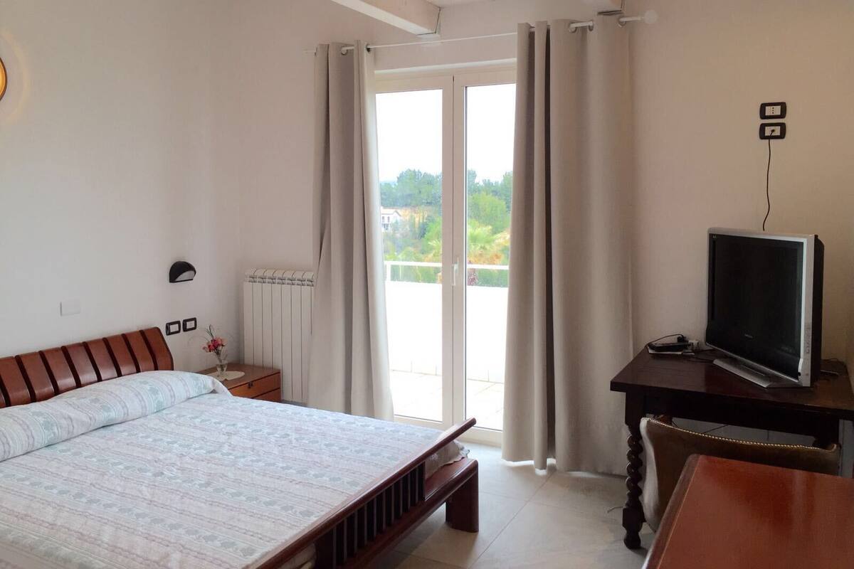 Une chambre à coucher avec un lit, un bureau avec une télévision, un radiateur et une fenêtre avec des rideaux.