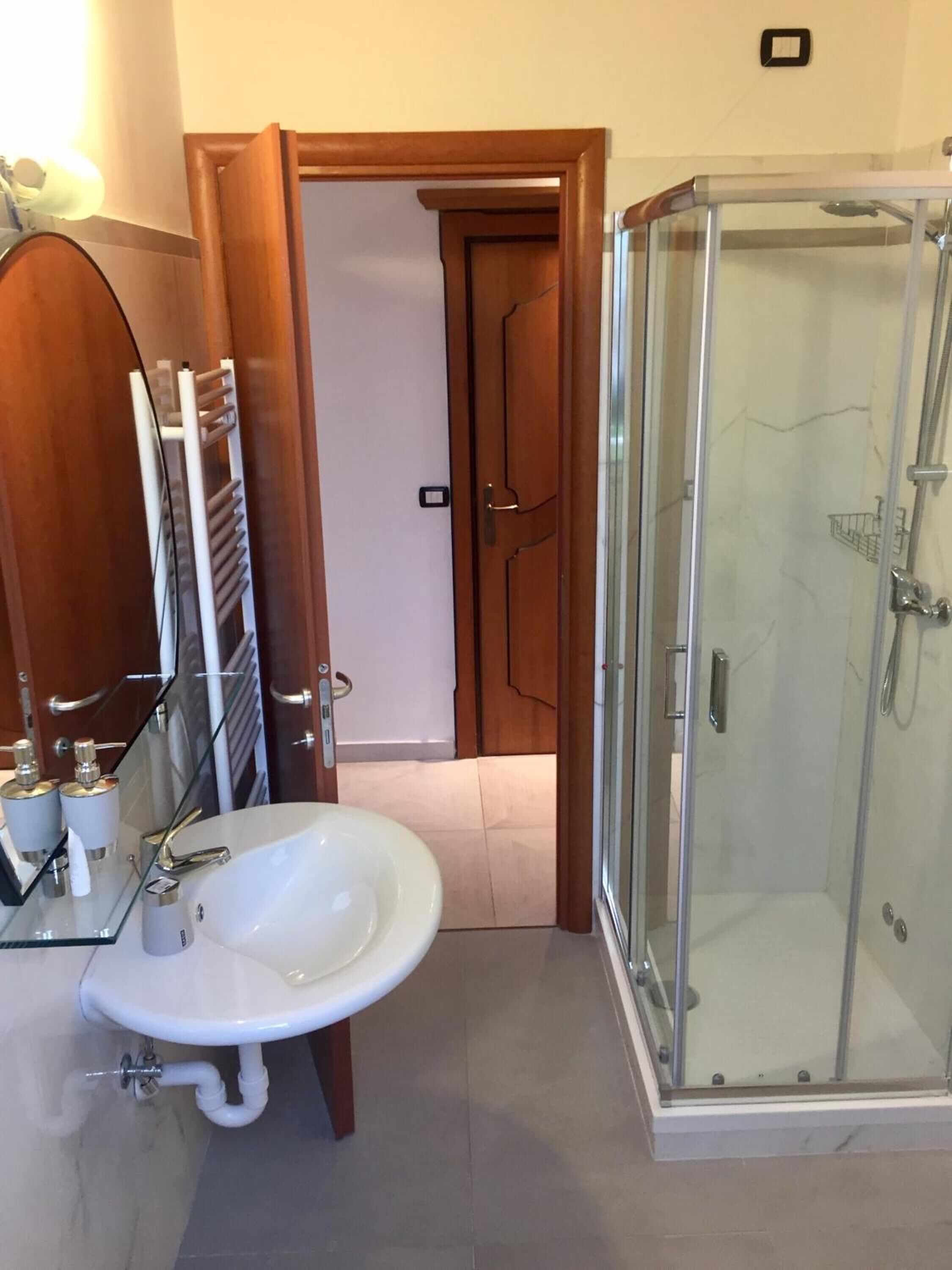 A bathroom with a glass-enclosed shower, a white sink with a glass shelf, and a wooden door leading to another room.