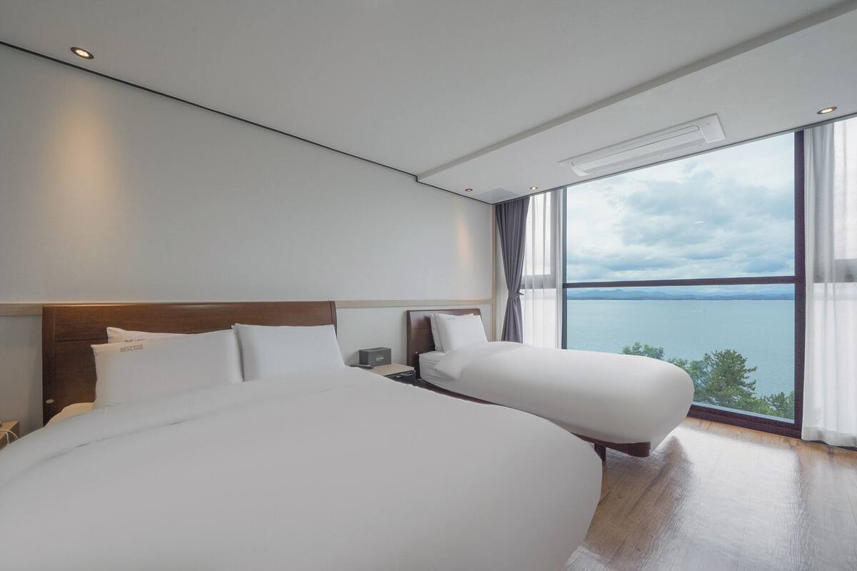 A modern hotel room with two large beds, a wooden headboard, a nightstand, and a large window offering a scenic view of the sea.