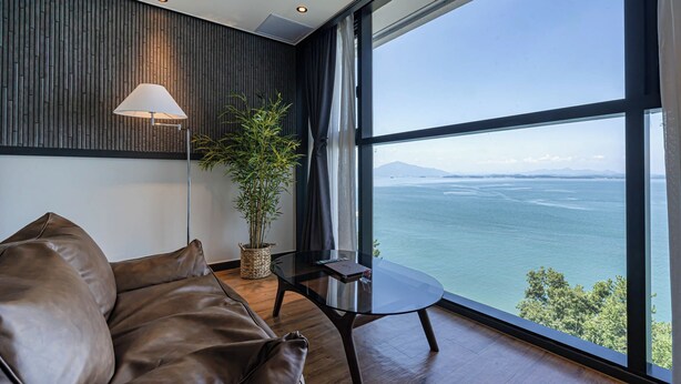 A modern living room with a leather sofa, a glass table, a floor lamp, and a large window offering a view of the sea and mountains. - Hotel Urban Island (Namhae)
