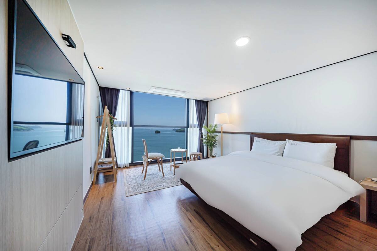 A modern hotel room with a large bed, a desk, and a view of the sea.