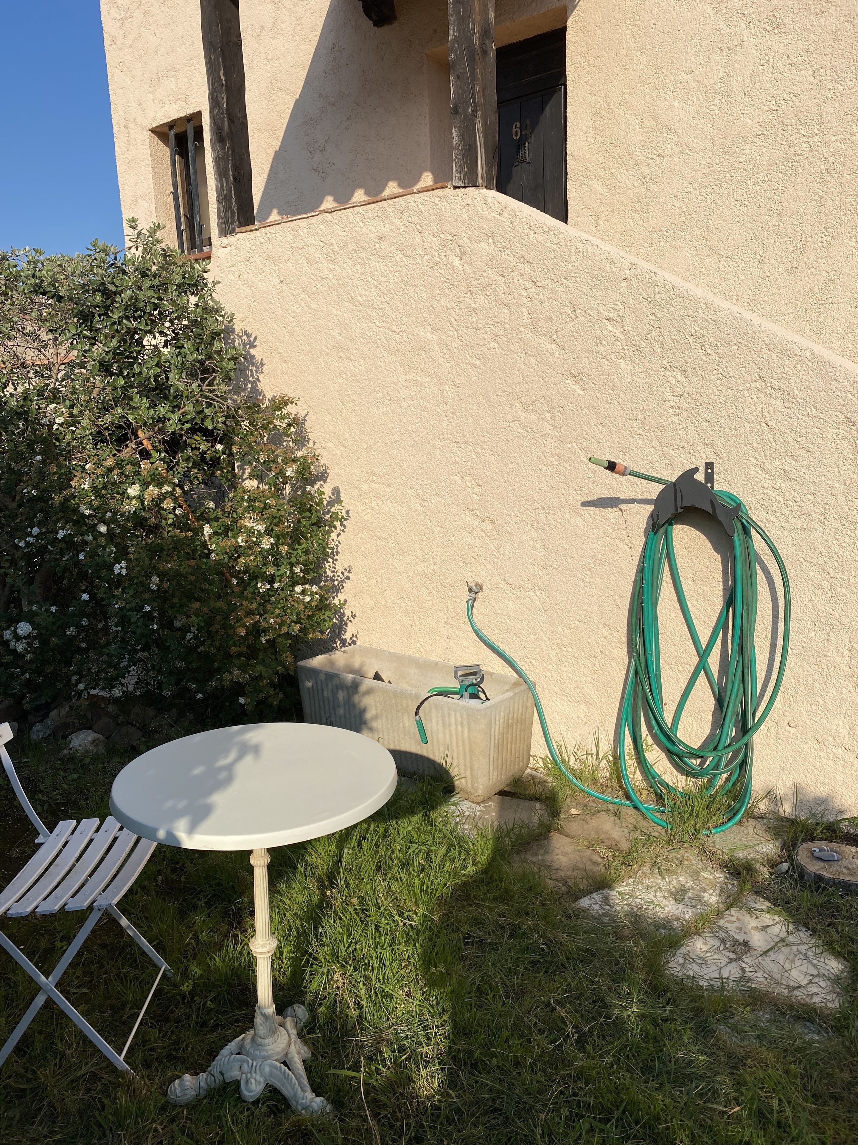 A concrete planter with a hose, a white table and chair, and a bush with white flowers.