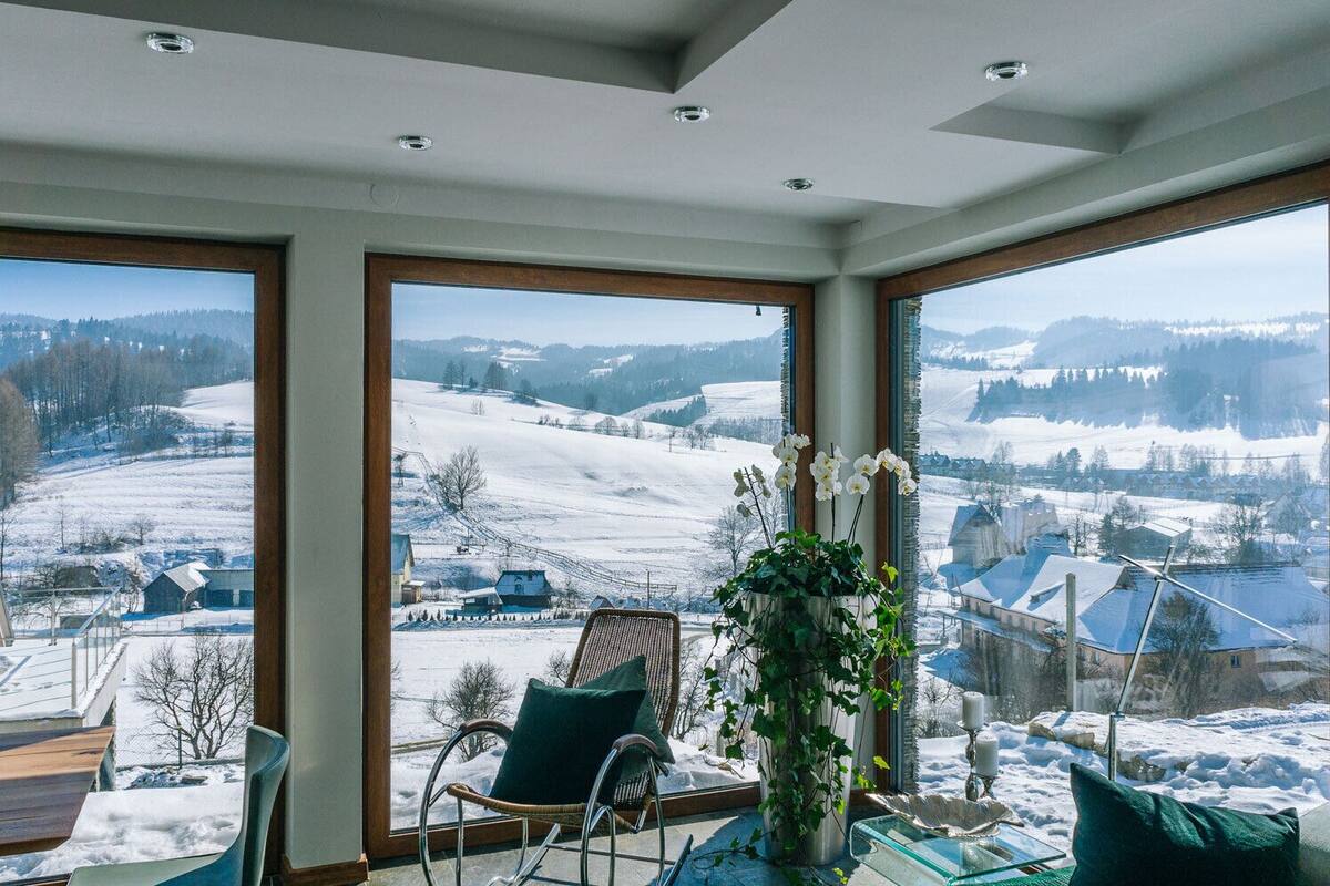 Ein Zimmer mit großen Fenstern und Blick auf eine verschneite Landschaft, ein Korbstuhl und ein Tisch mit Blumenvase.
