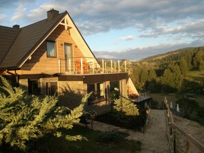 A modern house with a large balcony, stone facade, and a view of a forested valley.