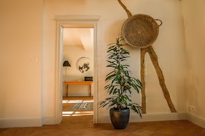 Un couloir avec une plante en pot, un panier tressé accroché au mur et une table en bois avec une lampe en arrière-plan.
