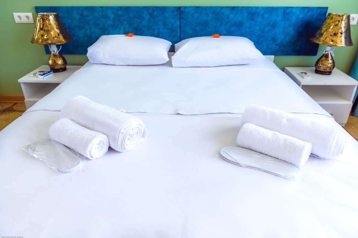 A neatly made bed with white linens, two pillows, and two rolled towels. There are two bedside tables with lamps and a wall socket.