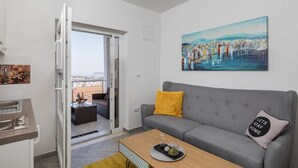 Una sala de estar moderna con un sofá gris, un cojín amarillo, un cuadro de un paisaje urbano y un balcón con vistas.