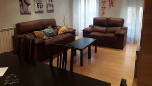 Sala de estar com dois sofás castanhos, uma mesa pequena e uma mesa de jantar com cadeiras. Há decorações nas paredes e uma janela com cortinas.
