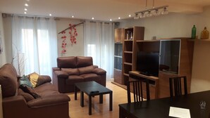 Uma sala de estar com um sofá de couro marrom, uma mesa de centro, uma TV sobre um suporte de madeira e uma mesa de jantar com cadeiras.