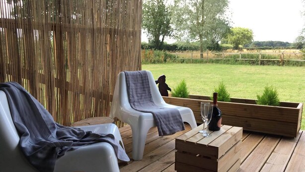 A wooden deck with two white plastic chairs, a wooden table with a bottle and glasses, and a wooden planter. - Wingfield Escapes (Diss)