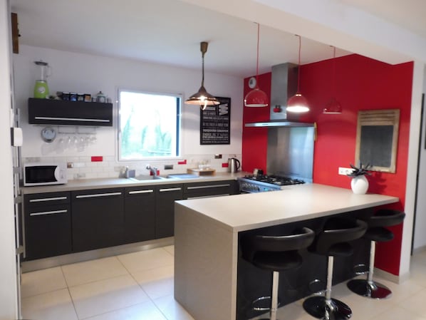 A modern kitchen with a central island, black bar stools, a red accent wall, and a window providing natural light.