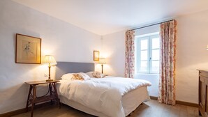 Ein Schlafzimmer mit einem Bett, zwei Nachttischlampen, einem hölzernen Nachttisch, einem gerahmten Kunstwerk an der Wand und einem Fenster mit blumigen Vorhängen.