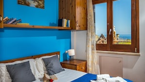 A bedroom with a bed, pillows, a bedside table with a lamp, a window with a view of a village, and a shelf with books. - Villa Elena 2 Km From the Natural Reserve of Zingaro (Castellammare del Golfo)