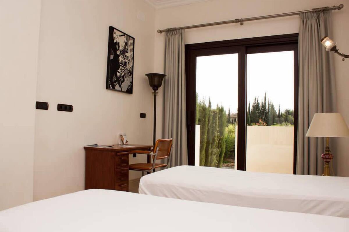 Habitación de hotel con dos camas, un escritorio, una silla, una lámpara y un ventanal grande con cortinas.