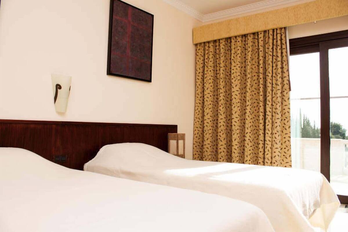 Habitación de hotel con dos camas, cabecero de madera, teléfono en la pared y una ventana con cortinas estampadas.