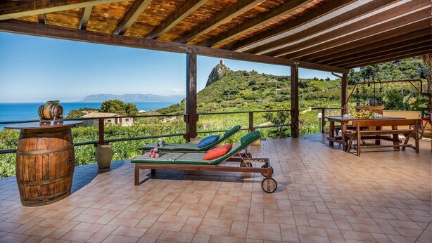 Ein überdachter Außenbereich mit Holzboden, einem Tisch mit Stühlen und Blick auf das Meer und die Hügel.
