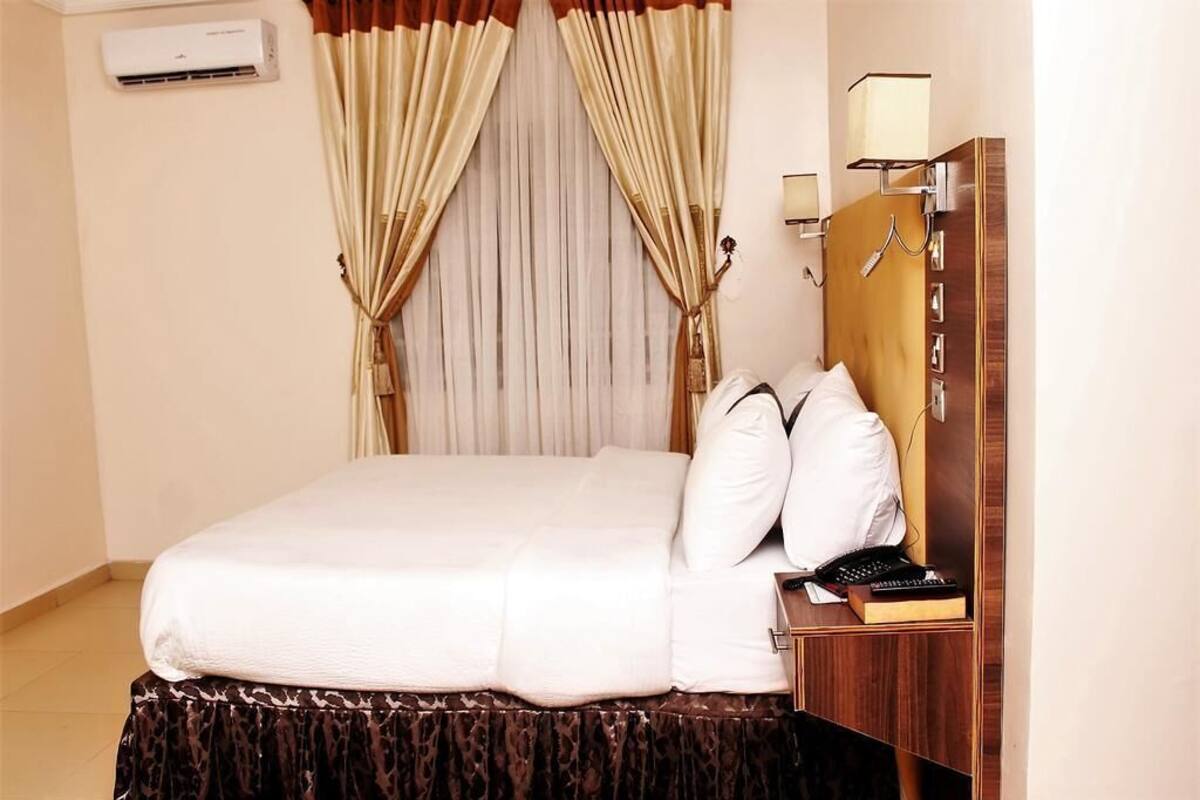 A hotel room with a bed, curtains, an air conditioning unit, a bedside table with a lamp, and a mirror.
