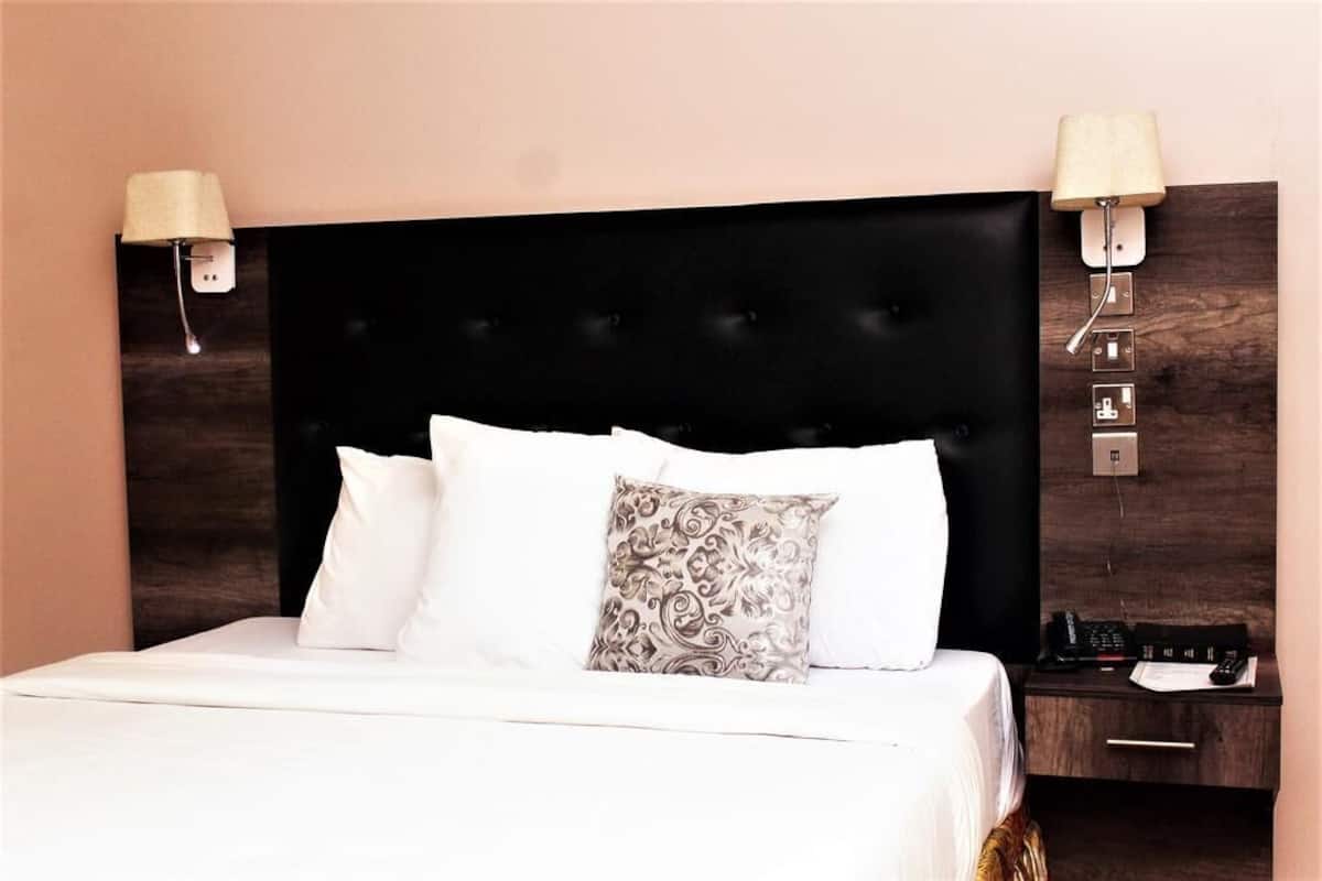 A hotel room with a black upholstered headboard, white bedding, a patterned throw pillow, bedside lamps, a nightstand with a phone and a remote control.