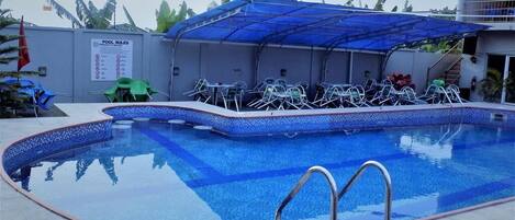 Una piscina con borde de azulejos azules, una zona de estar cubierta con sillas y un cartel con las normas de la piscina.