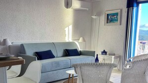 Un saló modern amb un sofà blau clar, cadires de vimet blanc, una taula petita i vistes al mar a través de grans finestres.