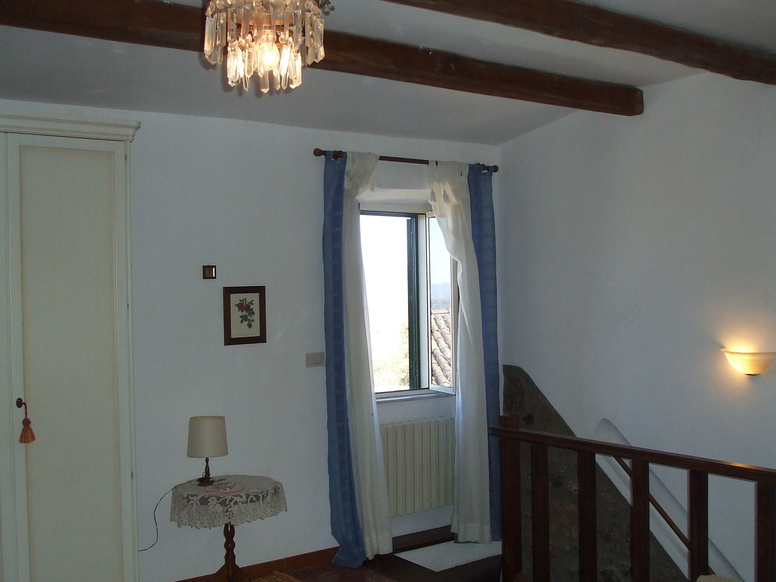 Una habitación con candelabro, una puerta, una pequeña mesa con lámpara, una ventana con cortinas y una escalera.