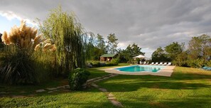 A swimming pool surrounded by a well-maintained lawn, with a house and a gazebo in the background.
