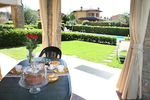 Un área de comedor al aire libre con una mesa preparada para una comida, un florero con flores rojas y vistas a una piscina y un jardín.
