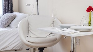 Une chambre d’hôtel moderne avec un lit blanc, une chaise blanche et une petite table à manger sur laquelle se trouvent un verre d’eau, une rose rouge et un verre à vin.