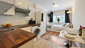 Une cuisine compacte équipée d’un lave-linge, un coin repas avec une table pour deux, et un lit avec vue sur la fenêtre.